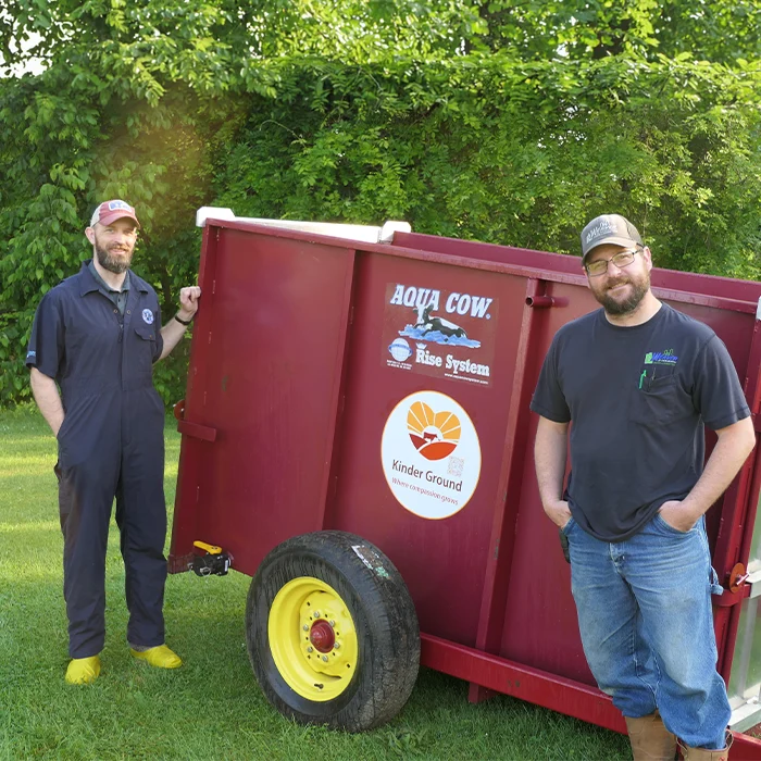 Two men stand beside a red Aqua Cow Rise System tank on grass, with trees in the background.