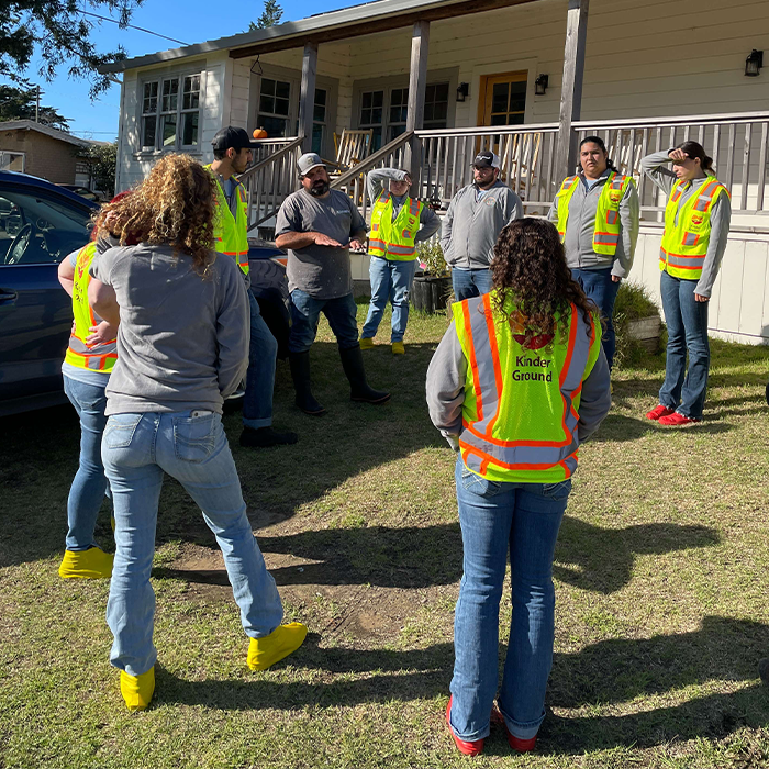 A group of workers in safety vests and yellow boots stand in a circle outside a house, listening to instructions.