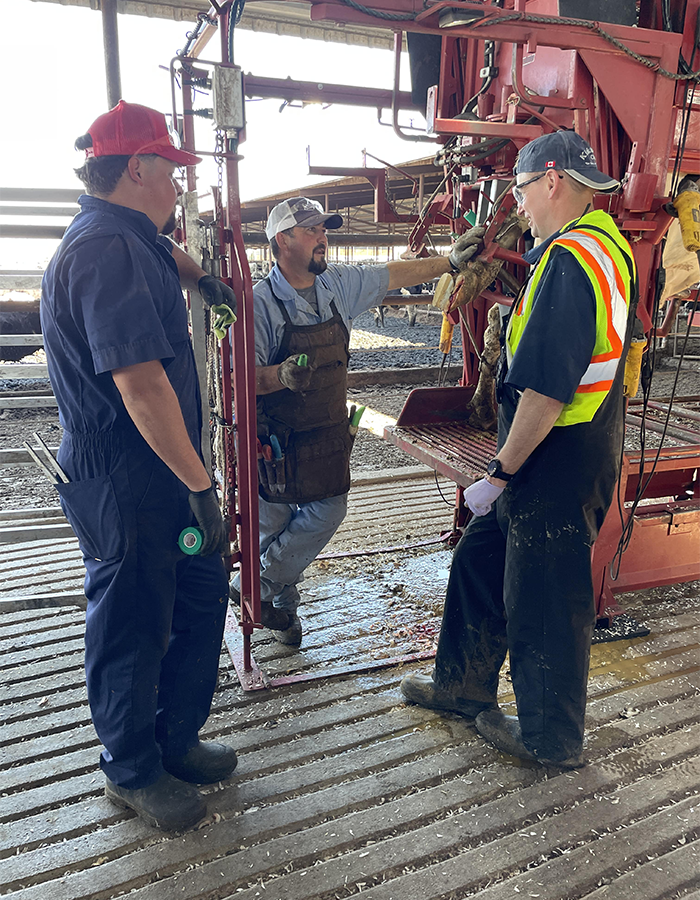 Three men in work clothes and safety gear talk next to red machinery in an industrial setting.