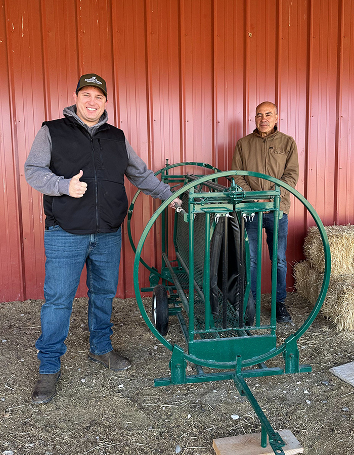 Two men stand by green livestock equipment in front of a red wall; one man is smiling and giving a thumbs up.