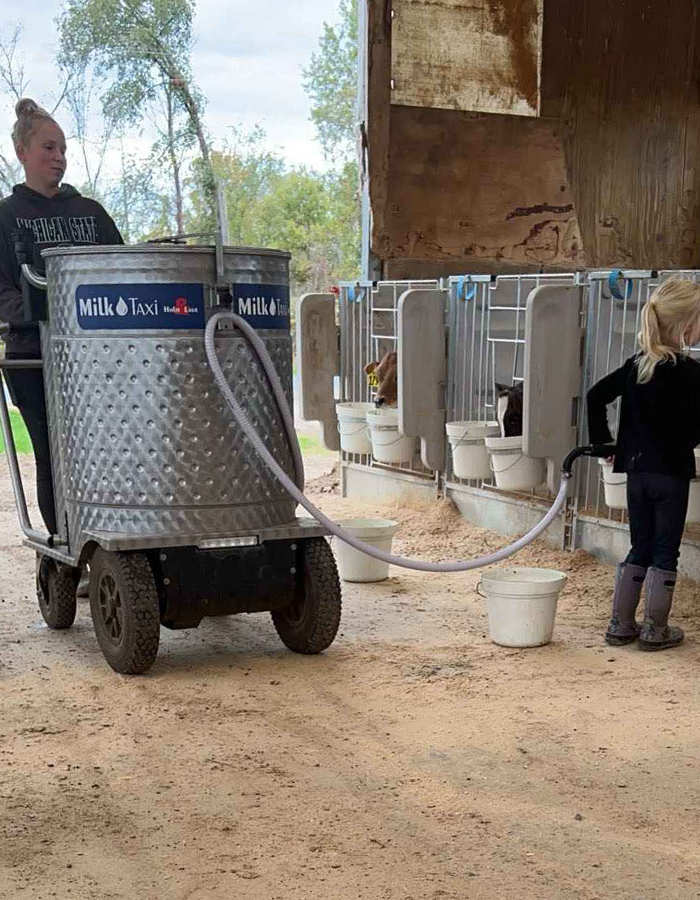 A woman uses a large Milk Taxi machine to fill buckets in a barn, with a child standing nearby.